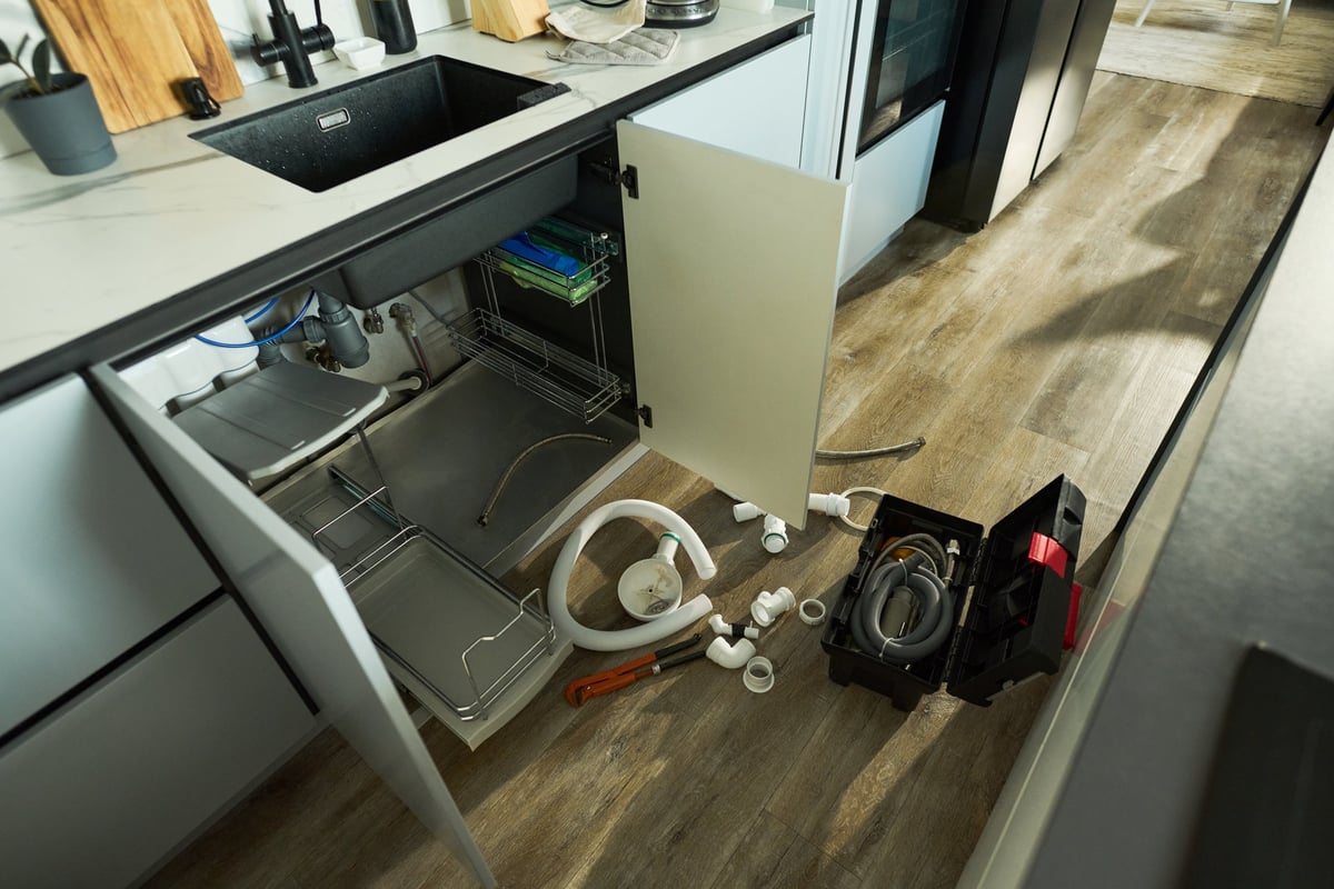 Kitchen scene showing open cabinet doors under sink with plumbing tools, pipes, and fittings scattered on floor, indicating ongoing repair or installation process, no people visible